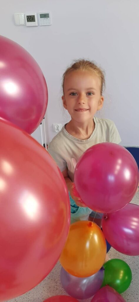 Uśmiechnięta dziewczynka patrząca w stronę fotografa zasłonięta za wyjątkiem twarzy napompowanymi balonami.
