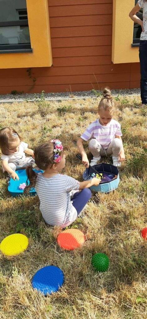Trzy dziewczynki siedzące na suchej trawie i bawiące się mini basenem dla lalek z zabawkami.